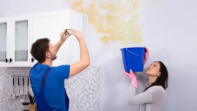 Homeowner photographing a water stained wall while a bucket catches a leak